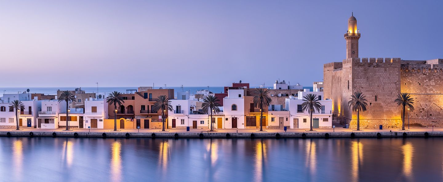 مدينة ساحلية بأسوار حصن قديم ومئذنة ونخيل على الميناء عند الغسق